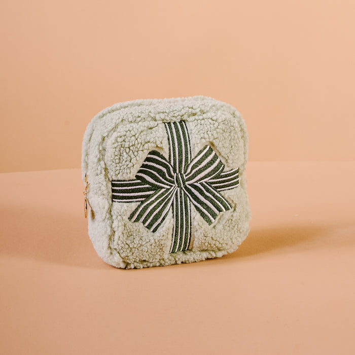 The Darling Effect’s Square Holiday Gift Bow Green Teddy Pouch is a fluffy, cream sherpa pouch with a zipper and a green and white striped bow, shown against a plain peach background.