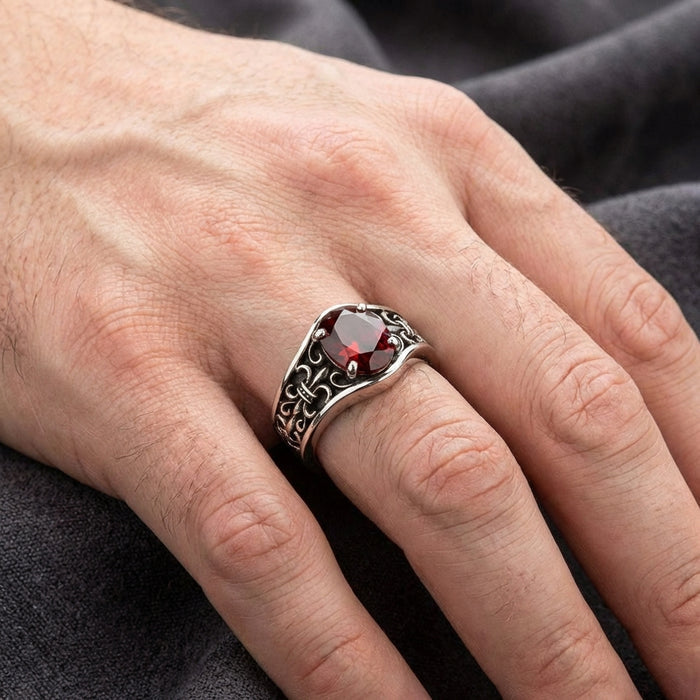 Sterling silver fleur-de-lis ring with deep red garnet gemstone worn on a man's ring finger, featuring ornate Gothic scrollwork design on the band against a dark gray fabric background