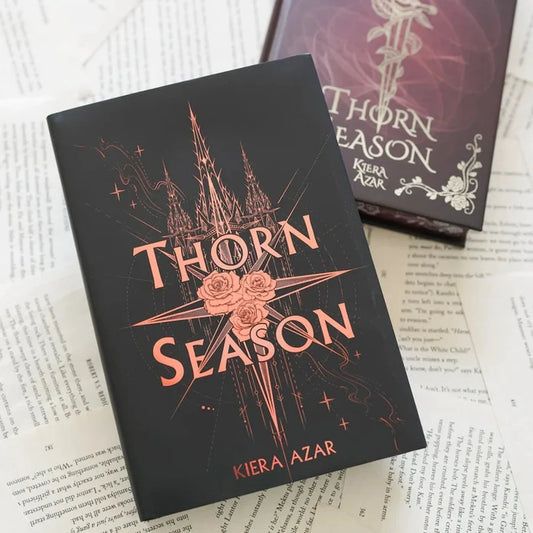 This image shows two books placed on top of what appears to be a stack of papers or documents. The book in the foreground has a black cover with the title "THORN SEASON" prominently displayed in large, stylized letters. The title is accompanied by an intricate design featuring a castle, roses, and other decorative elements. The author's name, "KIERA AZAR," is listed at the bottom of the cover. The book in the background has a similar design theme but with a different color scheme, featuring purp