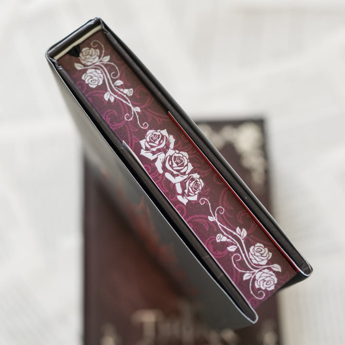 This image shows a close-up view of a book's top page edges with a maroon background and white floral designs, specifically roses.  The floral pattern is intricate, with detailed rose illustrations that add an elegant touch to the book. The book is placed on top of another object, which is partially visible and seems to have a similar design theme.