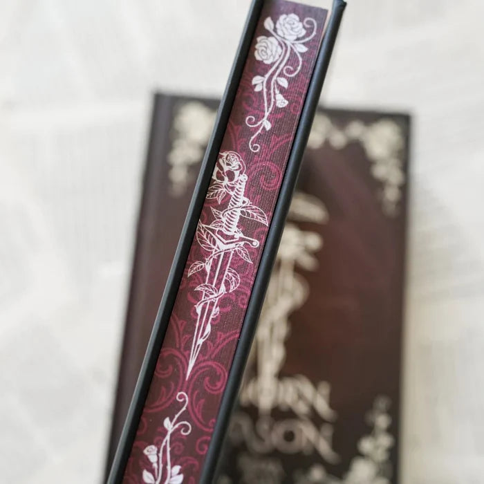 This image shows a close-up view of a book's page edges with a decorative design. The page edges feature a dark maroon background with a prominent white design that includes a sword and roses. The word "ASON" appears twice on the spine, suggesting it might be part of the book's title or author's name. The book appears to be placed on a surface with a soft, neutral background, possibly a table or a bed. The focus of the image is on the intricate design and text on the book page edges