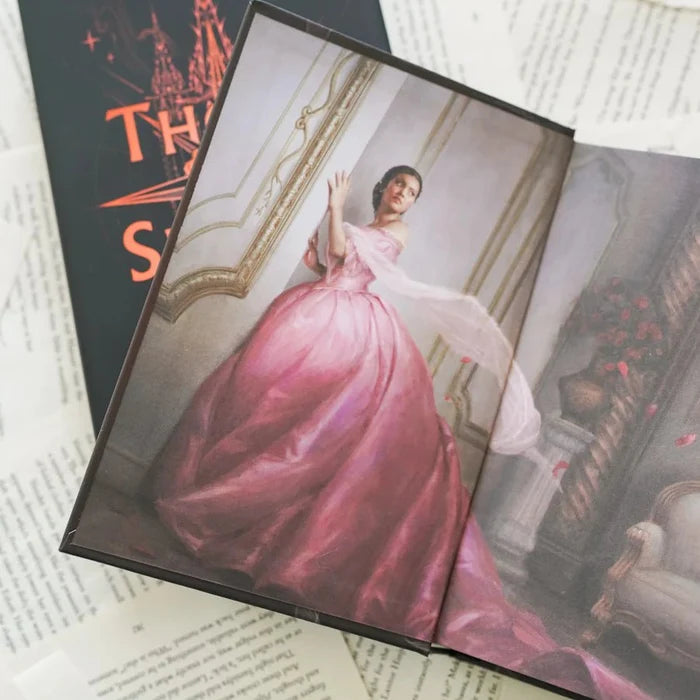 This image shows an open book displaying a detailed illustration of a woman in a lavish pink gown. The gown features a high neckline and long sleeves, with a flowing train and a white shawl draped over her arm. The woman is standing in an ornate room with classical decor, including a grand mirror and a chair. The book is placed on top of several sheets of paper with printed text, which appears to be part of a larger document or manuscript.