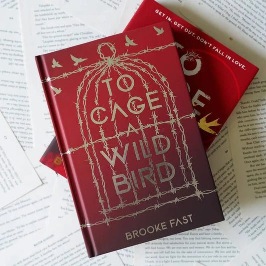 To Cage a Wild Bird LitJoy Edition by Brooke Fast — dark red hardcover with gold foil barbed wire birdcage and flying birds design, shown front and back on scattered open book pages flatlay
