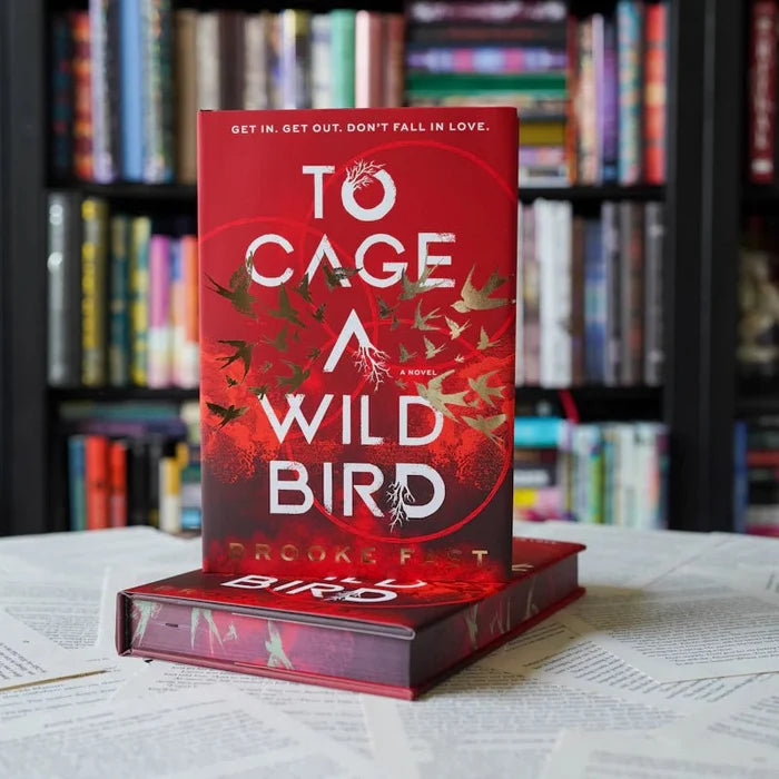 To Cage a Wild Bird by Brooke Fast — red hardcover with white title text and gold bird silhouettes spiraling on the cover, tagline "Get In. Get Out. Don't Fall in Love.," standing upright on another copy with a colorful bookshelf background