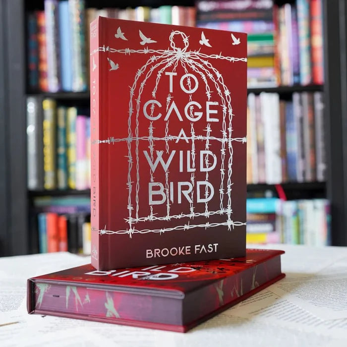To Cage a Wild Bird LitJoy Edition by Brooke Fast — deep red hardcover with silver barbed wire birdcage and birds design, standing upright on another copy with a colorful blurred bookshelf background