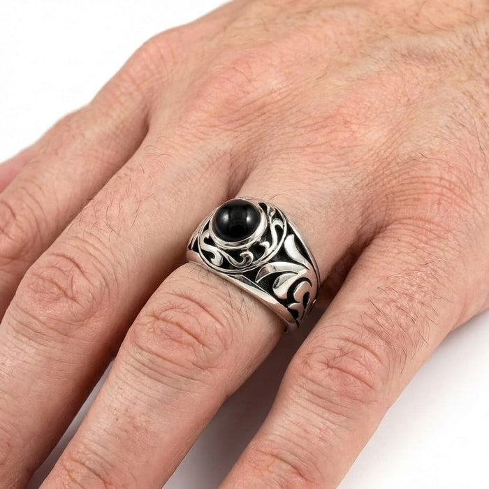 Close-up of a man's hand wearing a vintage-style silver men's ring featuring a round black onyx gemstone and intricate filigree swirl detailing against a white background.