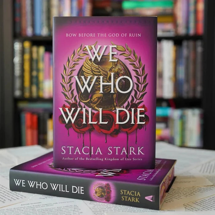 We Who Will Die LitJoy Edition hardcover by Stacia Stark — purple cover with gold laurel wreath, winged figure, and red roses, stacked on book pages with bookshelf background, from LitJoy Winter Romantasy TBR Box