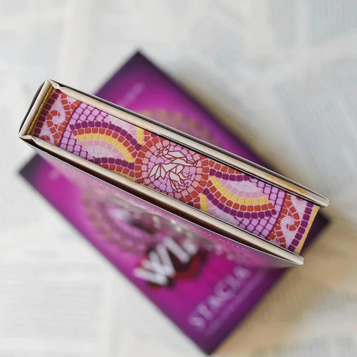 Overhead close-up of We Who Will Die LitJoy Edition lying flat, showing the top edge and mosaic-patterned cover detail in pink, orange, and gold tile design, resting on the purple standard edition