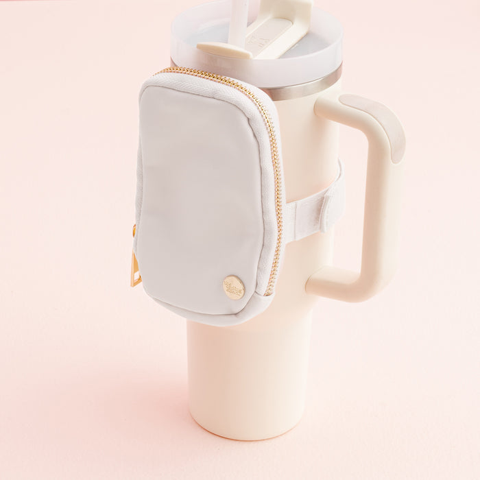 A White Tumbler Fanny Pack by The Darling Effect is attached to a cream-colored travel mug with a handle and straw, shown on a pale pink surface.
