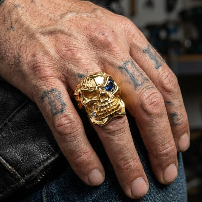 Yellow gold skull ring with blue CZ eye worn on tattooed hand showing detailed craftsmanship and substantial size