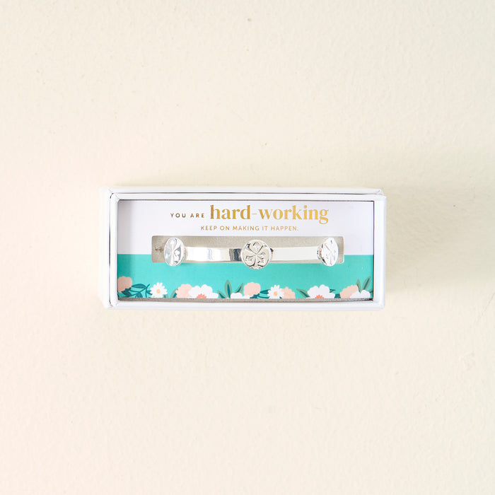The Darling Effect's You Are Hard-Working Skinny Stacker Bracelet, a silver adjustable cuff with engraved designs, is shown in a white gift box featuring the message "You are hard-working. Keep on making it happen." on a light cream background.